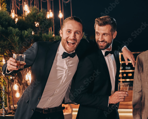 Two handsome men in suits drinking whiskey and smiling while spending time on party photo