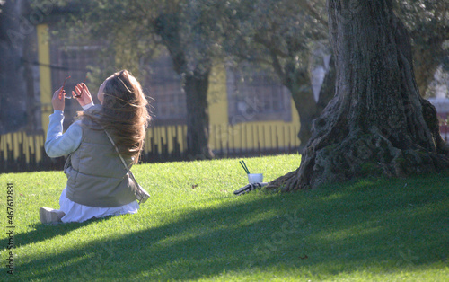 Rapariga sentada num jardim de cidade ao lado de uma árvore oliveira movimentar a cabeça para ajeitar o cabelo e a colocar os óculos - outono - descanso - apanhar sol - copo de refresco ao lado photo