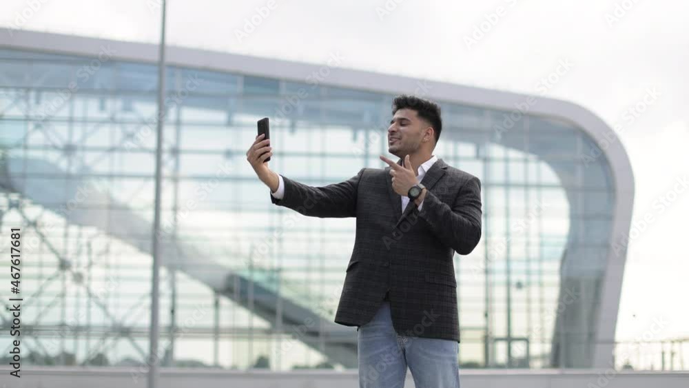 People, lifestyle, travel, tourism and modern technology. Handsome happy Indian businessman making video call on smartphone to his friend and showing hello gesture against modern airport background