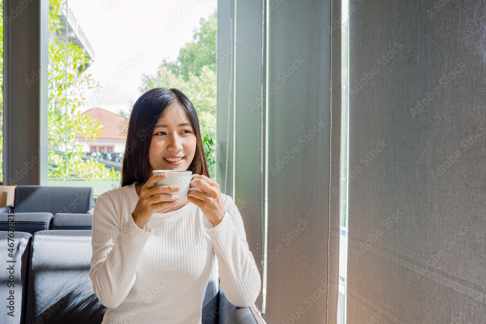 Beautiful asian woman drink a cup of coffee and tea in living room with curtain window. Happy asian girl relax with green nature through the glass window in home. Beautiful asian woman drink a cup of coffee and tea in living room with curtain window. Happy asian girl relax with green nature through the glass window in home.