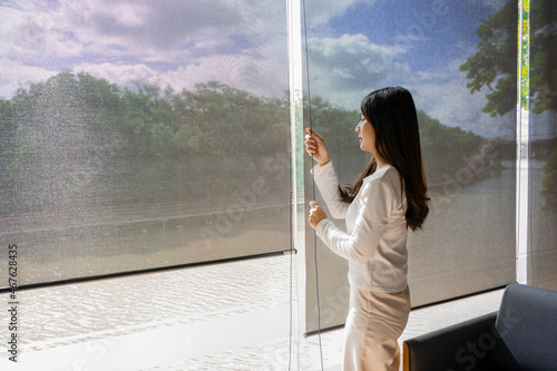 Happy asian woman relax with green nature through the glass window in home. Asian girl open the curtain blinds to looking outside and enjoy with sunlight in the morning.