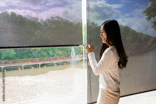Happy asian woman relax with green nature through the glass window in home. Asian girl open the curtain blinds to looking outside and enjoy with sunlight in the morning.