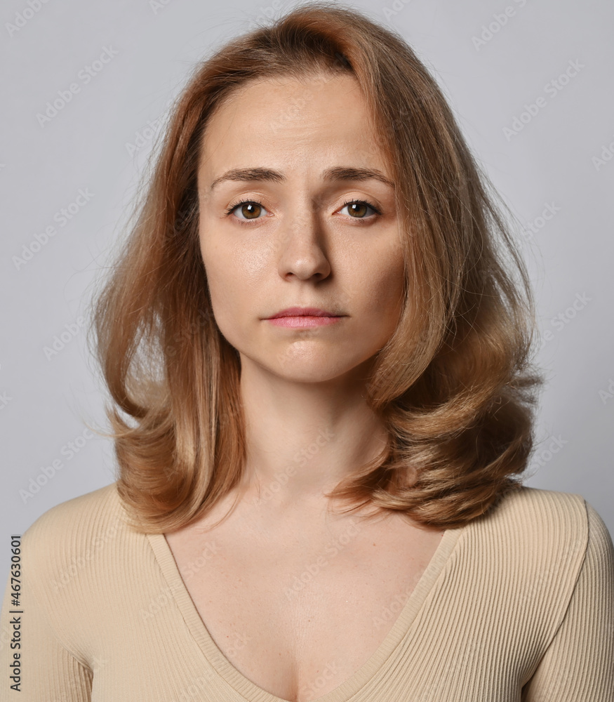 Fototapeta premium Portrait of young woman in beige shirt with long sleeves and volumetric blonde curly hair, with no smile looking at camera