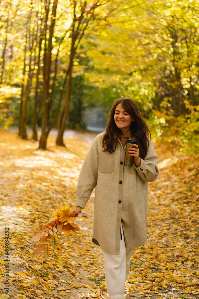 A young woman in a long shirt with a thermo mug in her hands walks through the autumn forest. Natural landscape. Beautiful forest. Autumn day. A young woman in a long shirt with a thermo mug in her hands walks through the autumn forest. Natural landscape. Beautiful forest. Autumn day.