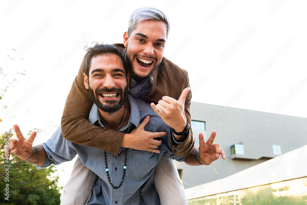 Playful smiling male gay couple looking at camera. Man piggyback ride with boyfriend. Copy space.