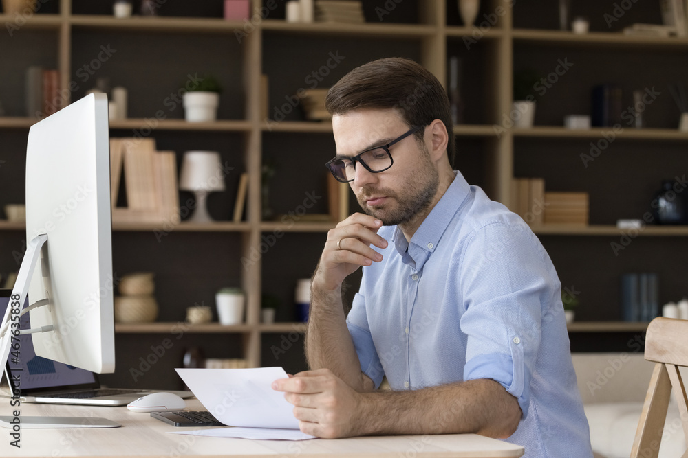 Focused skilled millennial businessman in eyewear looking through paper document, analyzing marketing research report or contract deal agreement, working on computer alone in modern office room. Focused skilled millennial businessman in eyewear looking through paper document, analyzing marketing research report or contract deal agreement, working on computer alone in modern office room.