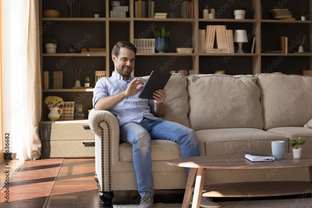 Happy young relaxed man using digital touchpad gadget, enjoying web surfing information, typing message communicating with friends, choosing goods shopping in internet store, sitting on cozy sofa. Happy young relaxed man using digital touchpad gadget, enjoying web surfing information, typing message communicating with friends, choosing goods shopping in internet store, sitting on cozy sofa.