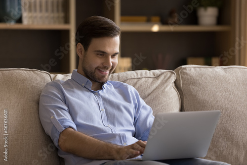 Happy young man working distantly on computer at home, sitting on comfortable sofa. Smiling millennial handsome guy using laptop, web surfing information, communicating distantly or shopping online.