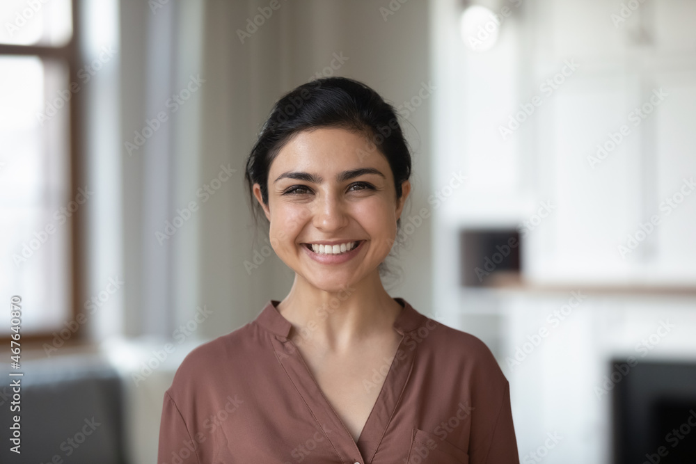Head shot attractive young 30s Indian female with wide toothy smile posing on camera while standing alone in modern cozy living room at own or rented apartment. Tenancy, housekeeper portrait concept - obrazy, fototapety, plakaty 