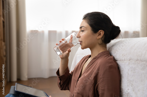 Side profile attractive young Indian woman put digital tablet on laps distracted from modern tech usage  hold glass drink still water sit on floor leaned on bed. Healthy life habit  lifestyle concept