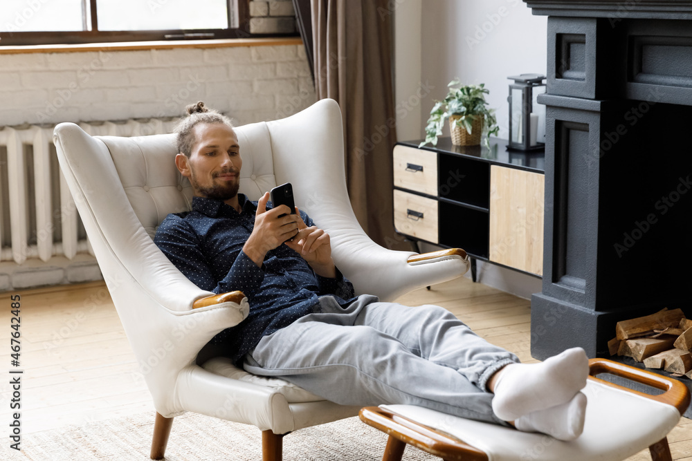 Happy relaxed young hipster man using cellphone applications, web surfing information, playing mobile games or spending time online in social networks, sitting in cozy armchair with legs on footstool.