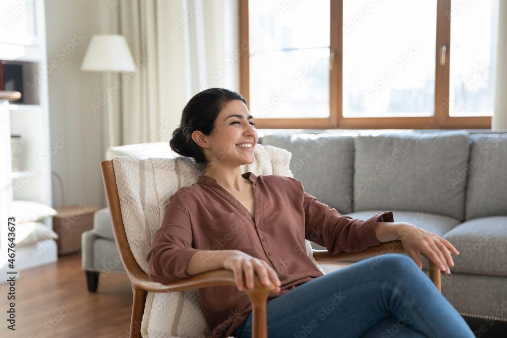 Happy Indian ethnicity woman relaxing on comfy wooden armchair spend time in cozy living room, smile enjoy carefree weekend and comfort furniture, breath fresh conditioned air inside modern apartment