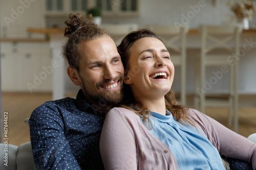 Happy affectionate young family couple man woman resting on cozy sofa, having fun watching smart television programs, entertaining enjoying leisure weekend time, daydreaming together at home.