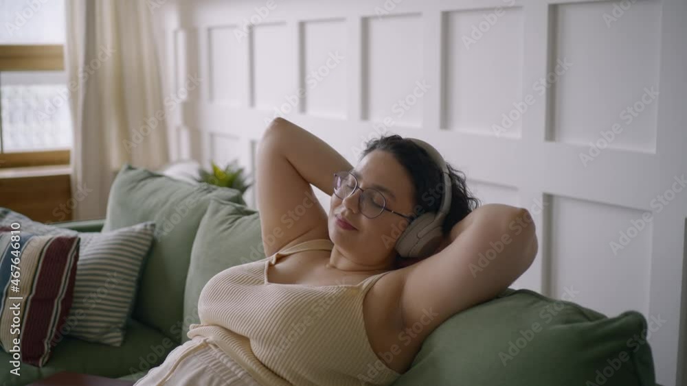 Curly haired overweight young woman wearing comfortable clothes lies on green sofa listening to music in wireless headphones in stylish room Curly haired overweight young woman wearing comfortable clothes lies on green sofa listening to music in wireless headphones in stylish room