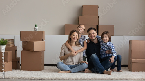 Portrait of happy family with kids enjoying moving day, hugging, sitting on floor with cardboard boxes, looking at camera, smiling mother and father with children posing for picture in new home