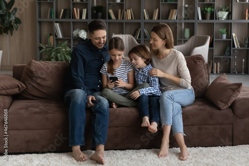 Happy family with kids using smartphone together, sitting on couch at home, smiling mother and father with little son and daughter having fun, looking at phone screen, enjoying leisure time