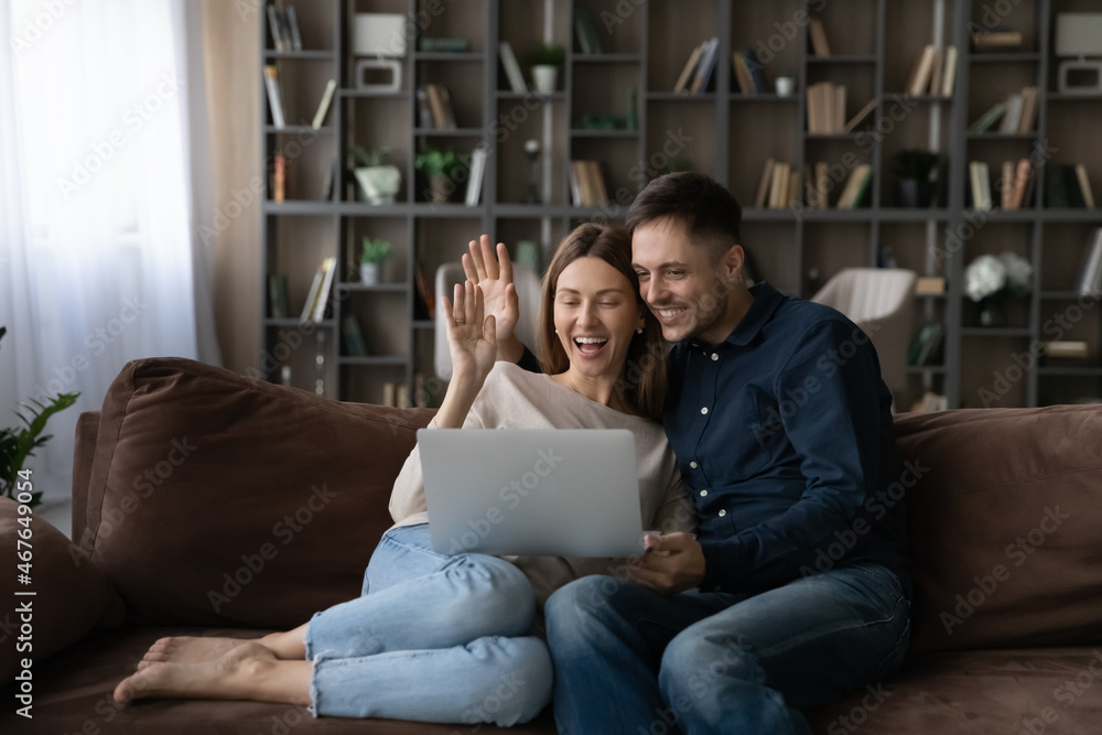 Happy couple hugging waving hands, chatting online with relatives or friends by video call, looking at laptop screen, sitting on couch at home, smiling wife and husband engaged in internet meeting Happy couple hugging waving hands, chatting online with relatives or friends by video call, looking at laptop screen, sitting on couch at home, smiling wife and husband engaged in internet meeting