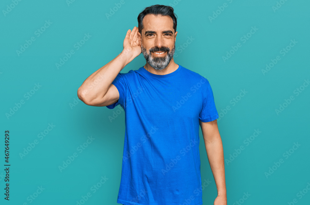 Middle aged man with beard wearing casual blue t shirt smiling with hand over ear listening an hearing to rumor or gossip. deafness concept.