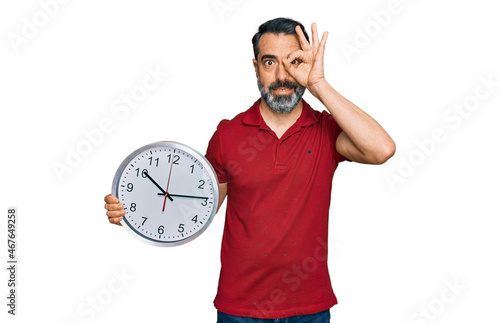 Middle aged man with beard holding big clock smiling happy doing ok sign with hand on eye looking through fingers