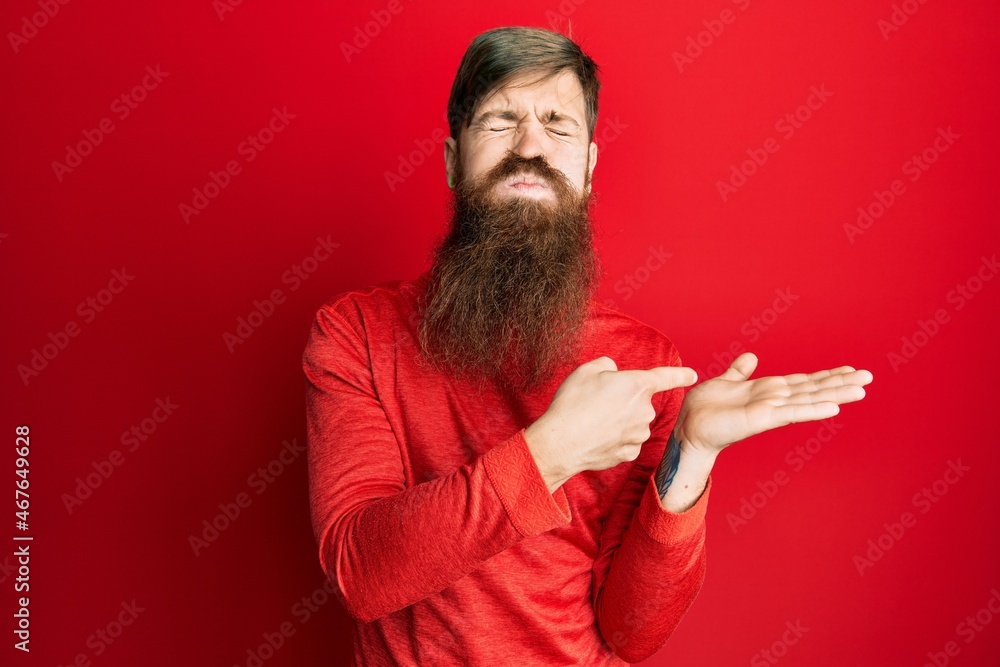 Fototapeta premium Redhead man with long beard pointing open palm with finger puffing cheeks with funny face. mouth inflated with air, catching air.