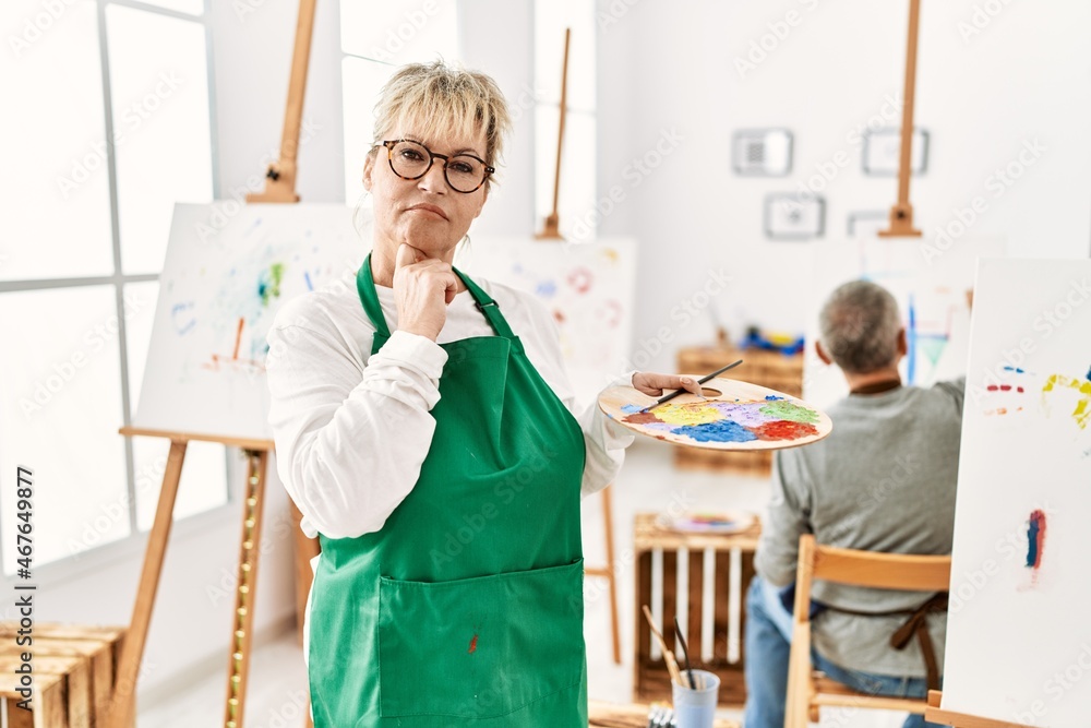Middle age caucasian woman drawing canvas at art studio serious face thinking about question with hand on chin, thoughtful about confusing idea