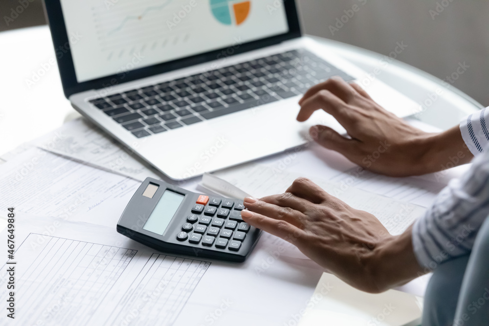 Cropped close up African American woman calculating bills or taxes, using laptop and calculator, businesswoman accountant working on financial report, checking documents, preparing presentation Cropped close up African American woman calculating bills or taxes, using laptop and calculator, businesswoman accountant working on financial report, checking documents, preparing presentation
