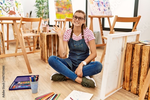Young brunette woman at art studio sitting on the floor showing and pointing up with fingers number five while smiling confident and happy.