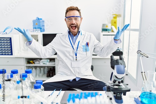 Middle age man working at scientist laboratory celebrating mad and crazy for success with arms raised and closed eyes screaming excited. winner concept