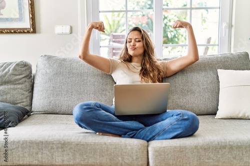 Beautiful young brunette woman sitting on the sofa using computer laptop at home showing arms muscles smiling proud. fitness concept.