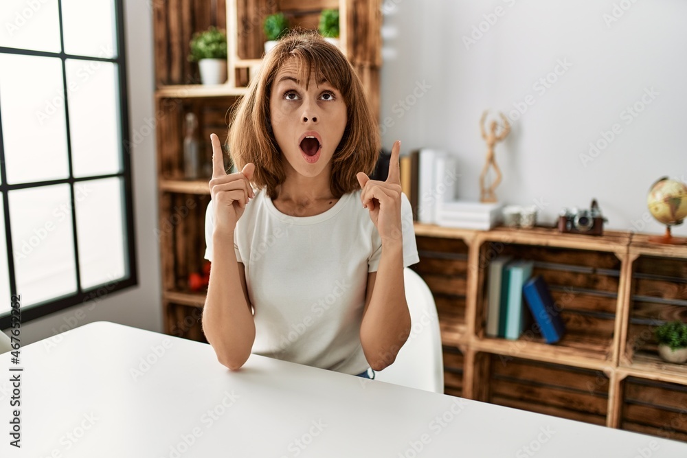Young caucasian girl wearing casual clothes sitting on the table at home amazed and surprised looking up and pointing with fingers and raised arms. Young caucasian girl wearing casual clothes sitting on the table at home amazed and surprised looking up and pointing with fingers and raised arms.