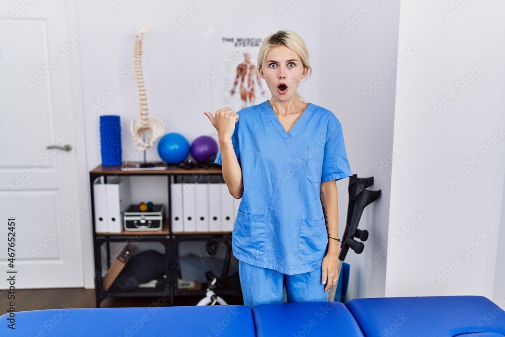 Young physiotherapist woman working at pain recovery clinic surprised pointing with hand finger to the side, open mouth amazed expression. Young physiotherapist woman working at pain recovery clinic surprised pointing with hand finger to the side, open mouth amazed expression.