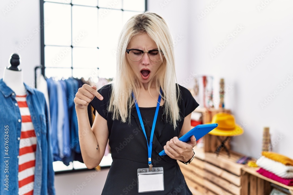 Young blonde woman working as manager at retail boutique pointing down with fingers showing advertisement, surprised face and open mouth Young blonde woman working as manager at retail boutique pointing down with fingers showing advertisement, surprised face and open mouth