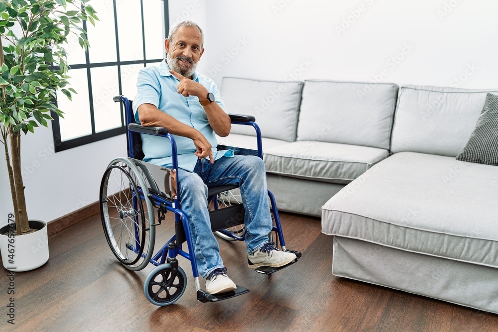 Handsome senior man sitting on wheelchair at the living room cheerful with a smile of face pointing with hand and finger up to the side with happy and natural expression on face Handsome senior man sitting on wheelchair at the living room cheerful with a smile of face pointing with hand and finger up to the side with happy and natural expression on face