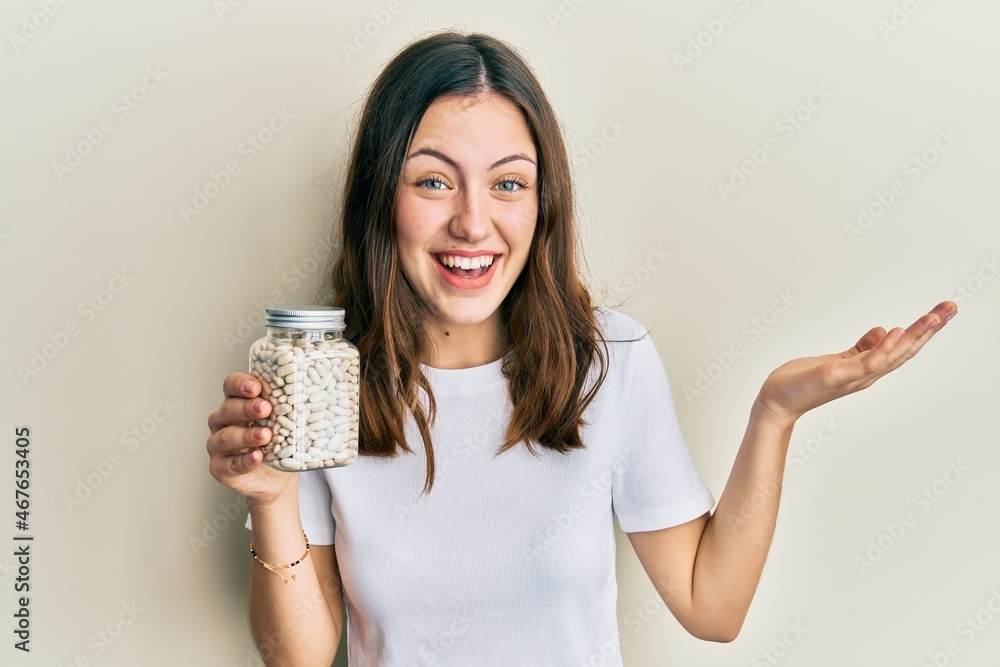 Young brunette woman holding bottle with raw white beans celebrating achievement with happy smile and winner expression with raised hand Young brunette woman holding bottle with raw white beans celebrating achievement with happy smile and winner expression with raised hand