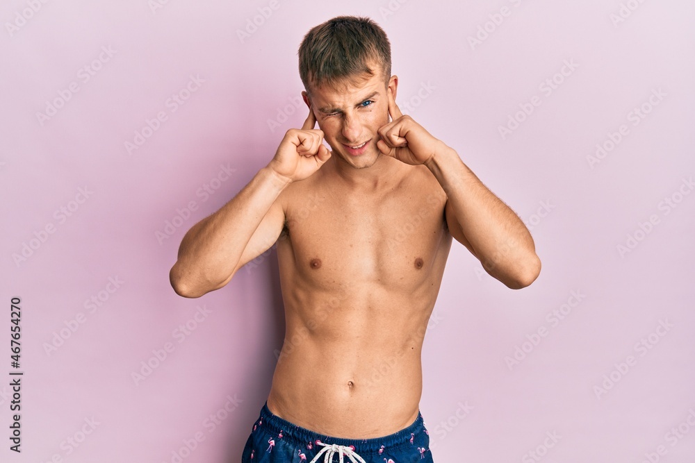 Fototapeta premium Young caucasian man wearing swimwear covering ears with fingers with annoyed expression for the noise of loud music. deaf concept.