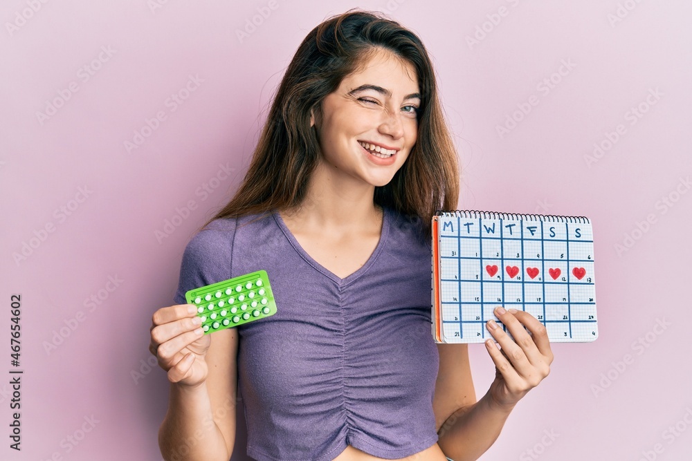Young caucasian woman holding birth control pills and menstrual calendar winking looking at the camera with sexy expression, cheerful and happy face. Young caucasian woman holding birth control pills and menstrual calendar winking looking at the camera with sexy expression, cheerful and happy face.