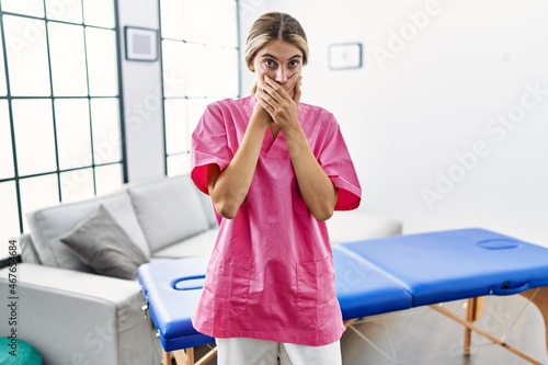 Young blonde woman working as physiotherapist at home shocked covering mouth with hands for mistake. secret concept.