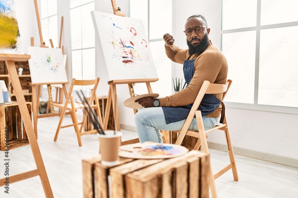 African american artist man painting on canvas at art studio with angry face, negative sign showing dislike with thumbs down, rejection concept