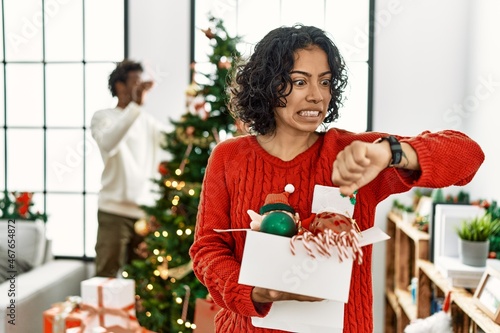 Young hispanic woman standing by christmas tree with decoration looking at the watch time worried, afraid of getting late