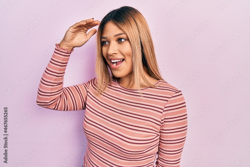 Fototapeta premium Beautiful hispanic woman wearing casual striped sweater very happy and smiling looking far away with hand over head. searching concept.