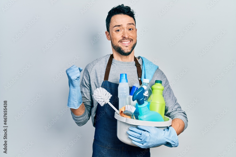 Young hispanic man holding cleaning products screaming proud, celebrating victory and success very excited with raised arm Young hispanic man holding cleaning products screaming proud, celebrating victory and success very excited with raised arm