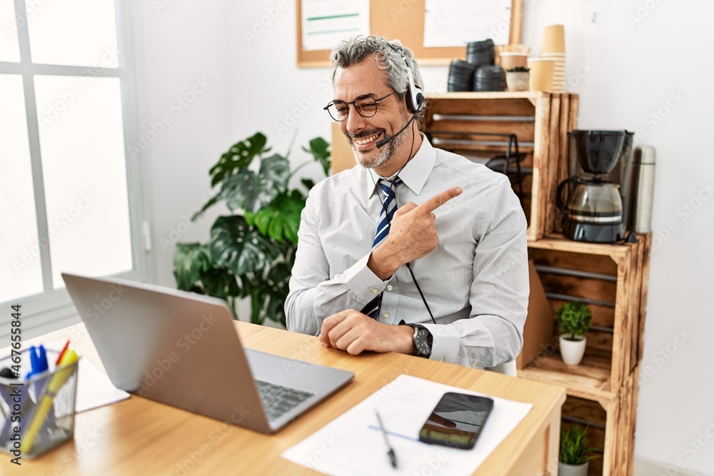 Middle age hispanic business man working at the office wearing operator headset cheerful with a smile of face pointing with hand and finger up to the side with happy and natural expression on face Middle age hispanic business man working at the office wearing operator headset cheerful with a smile of face pointing with hand and finger up to the side with happy and natural expression on face