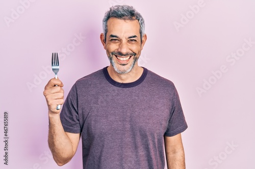 Handsome middle age man with grey hair holding one silver fork looking positive and happy standing and smiling with a confident smile showing teeth