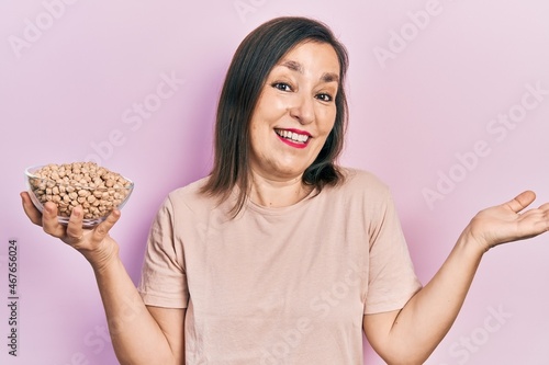 Middle age hispanic woman holding chickpeas bowl celebrating achievement with happy smile and winner expression with raised hand