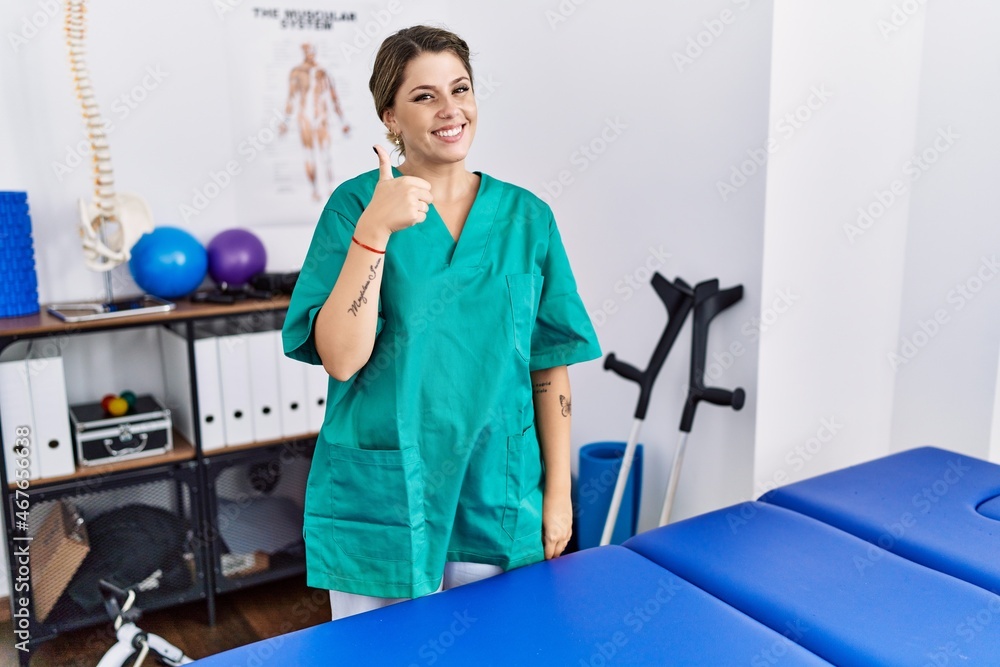 Young hispanic woman wearing physiotherapist uniform standing at clinic doing happy thumbs up gesture with hand. approving expression looking at the camera showing success. Young hispanic woman wearing physiotherapist uniform standing at clinic doing happy thumbs up gesture with hand. approving expression looking at the camera showing success.