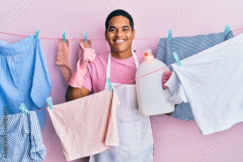 Young handsome hispanic man doing laundry holding detergent bottle smiling happy and positive, thumb up doing excellent and approval sign