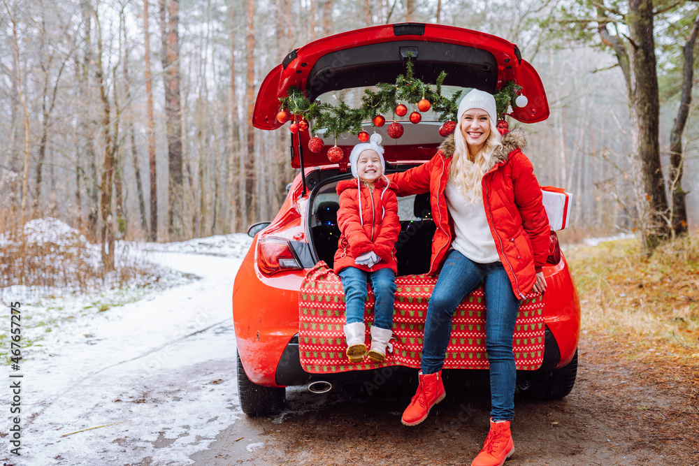 Happy family of blonde mom and little girl hug each other sitting in christmas decorated car in winter forest.