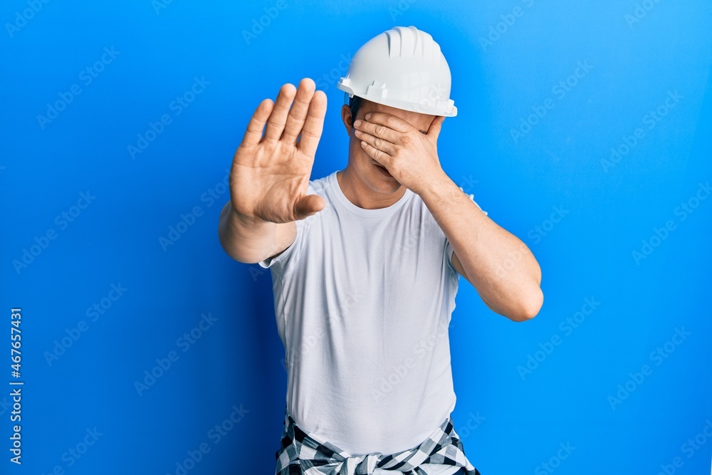 Handsome young man wearing builder uniform and hardhat covering eyes with hands and doing stop gesture with sad and fear expression. embarrassed and negative concept.