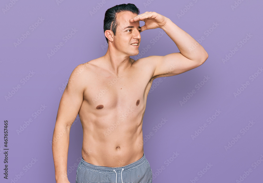 Handsome young man wearing swimwear shirtless very happy and smiling looking far away with hand over head. searching concept.
