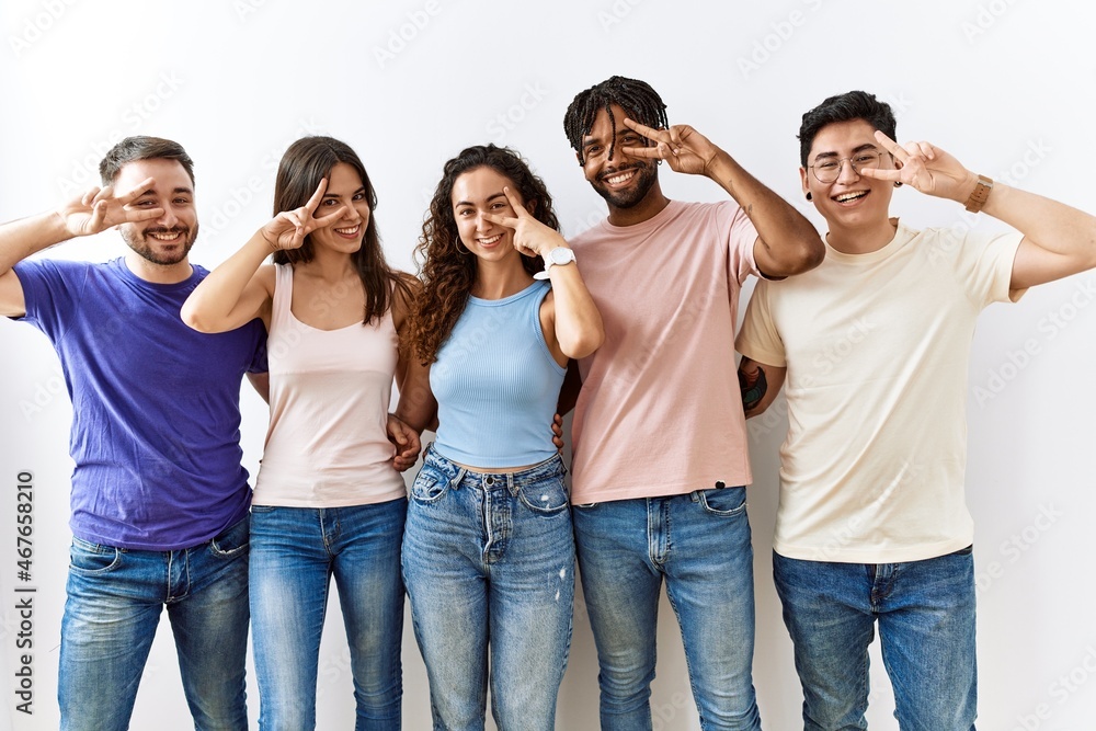 Group of young people standing together over isolated background doing peace symbol with fingers over face, smiling cheerful showing victory Group of young people standing together over isolated background doing peace symbol with fingers over face, smiling cheerful showing victory